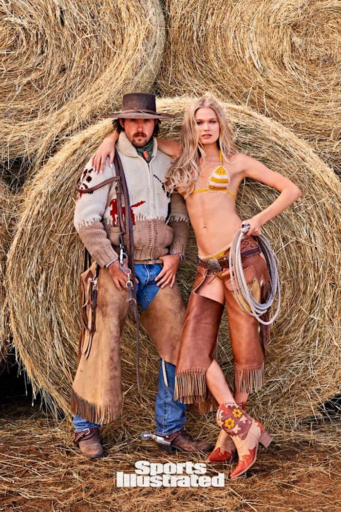 Vita Sidorkina stands in front of a hay bale in a knit bikini and brown leather chaps. She rests her arm on a man dressed in ranching garb.